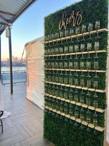 Image is of a green champagne wall with 6 rows of champagne glasses hanging on it. 