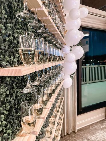 Image is of a green champagne wall with 6 rows of champagne glasses hanging on it. 