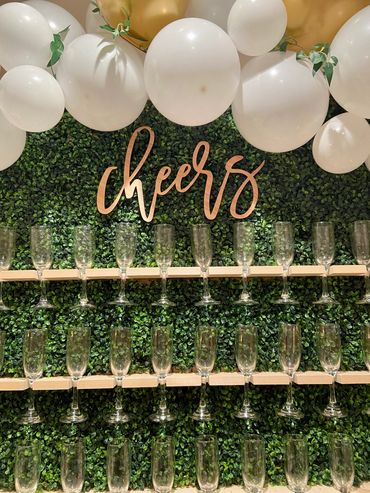 Image is of a green champagne wall with the word cheers in the center and balloons around it. 