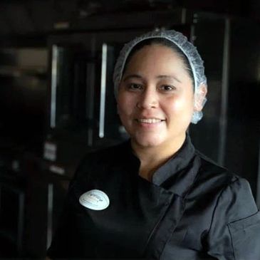 Smiling chef in black uniform and hairnet in a kitchen.