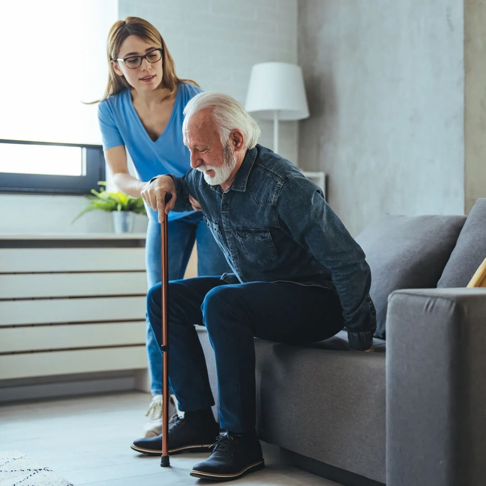 A young woman helps an elderly man stand up from a couch using a cane.