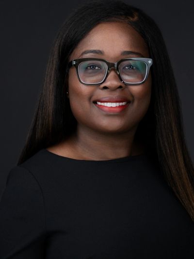 Professional portrait of a smiling woman with glasses and long hair against a black background.
