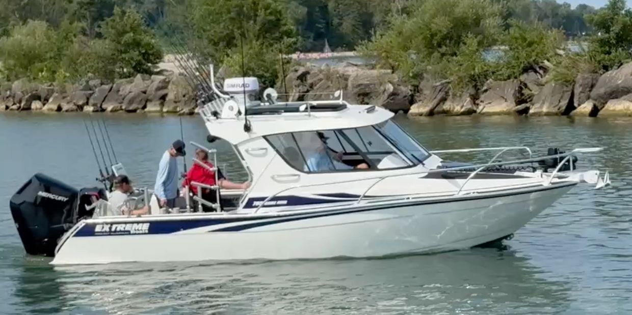 A white fishing boat with three people on a calm lake near rocks and trees.