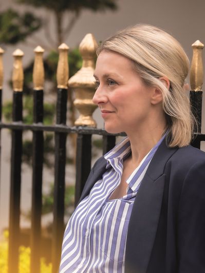 Woman in striped shirt and blazer stands outdoors near a decorative fence.