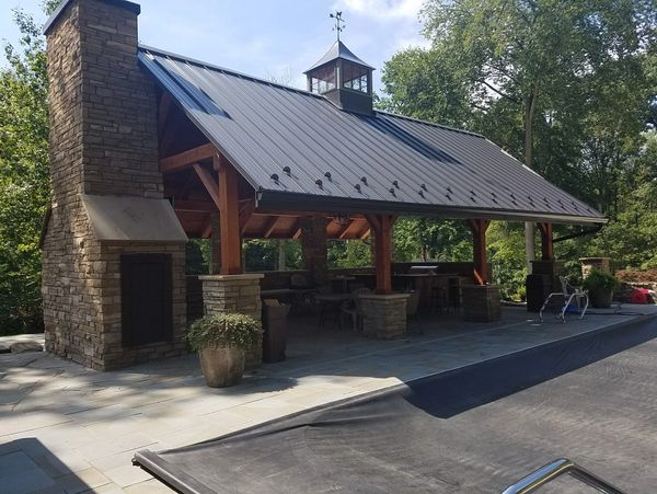 Outdoor pavilion with stone fireplace and metal roof near a pool.