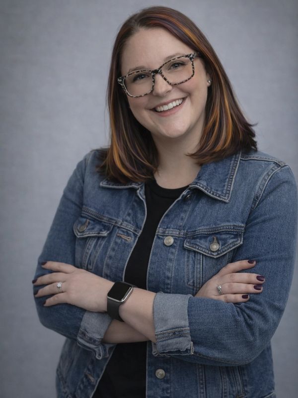 Smiling woman in glasses and denim jacket with arms crossed.