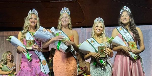 Four crowned beauty pageant winners holding flowers and plaques on stage.