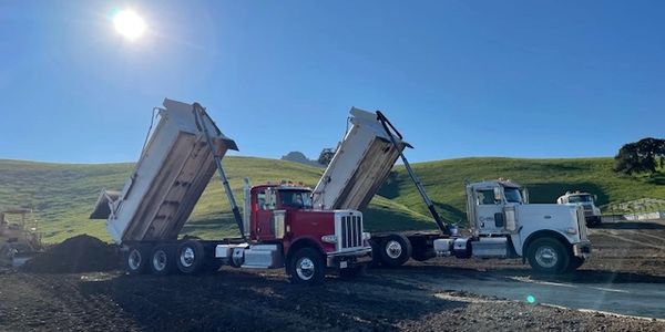 Two dump trucks unloading soil under a bright sun in a rural area.