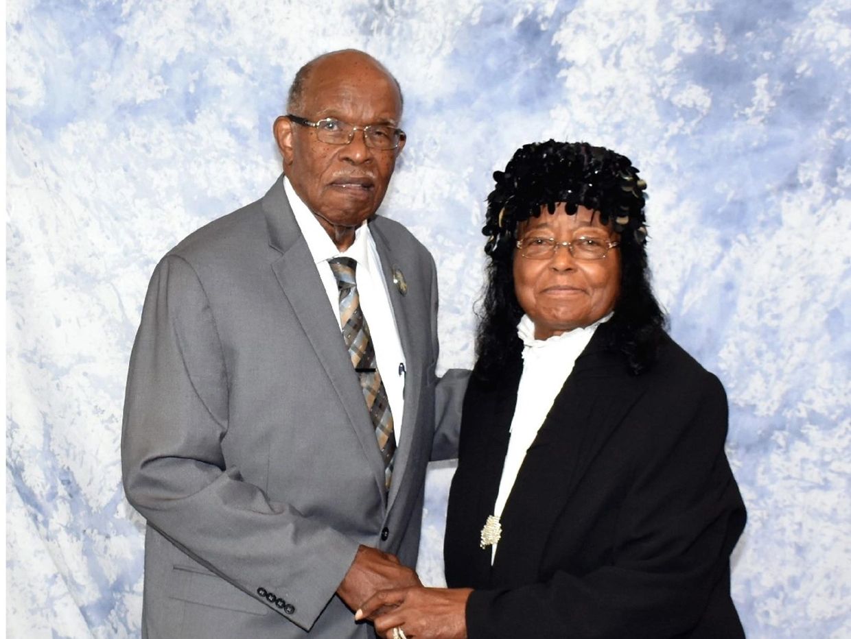 Elderly couple dressed formally, holding hands and posing together.