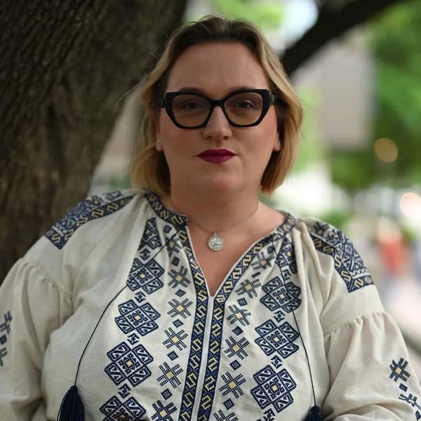 Woman with glasses and embroidered blouse standing outdoors.