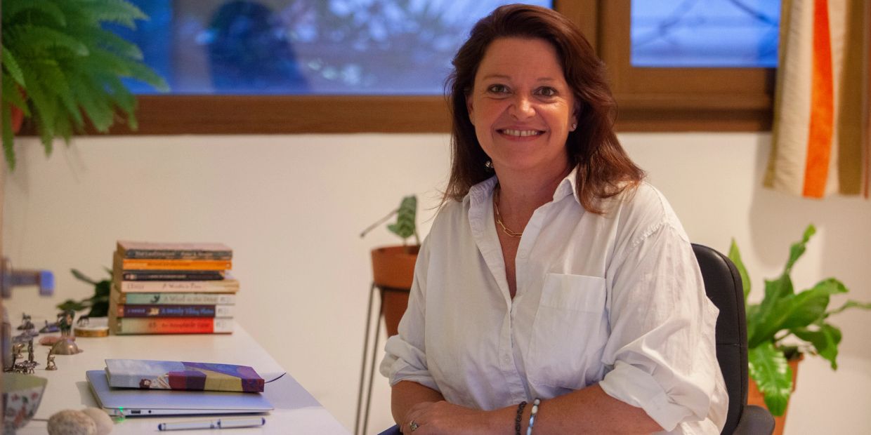 Smiling woman in white shirt sitting at a desk with books and plants.