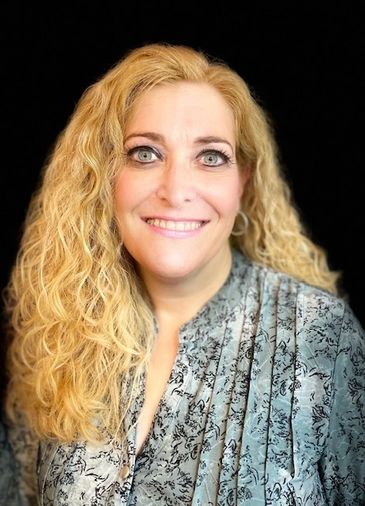Smiling woman with curly blonde hair wearing a patterned blouse against a black background.