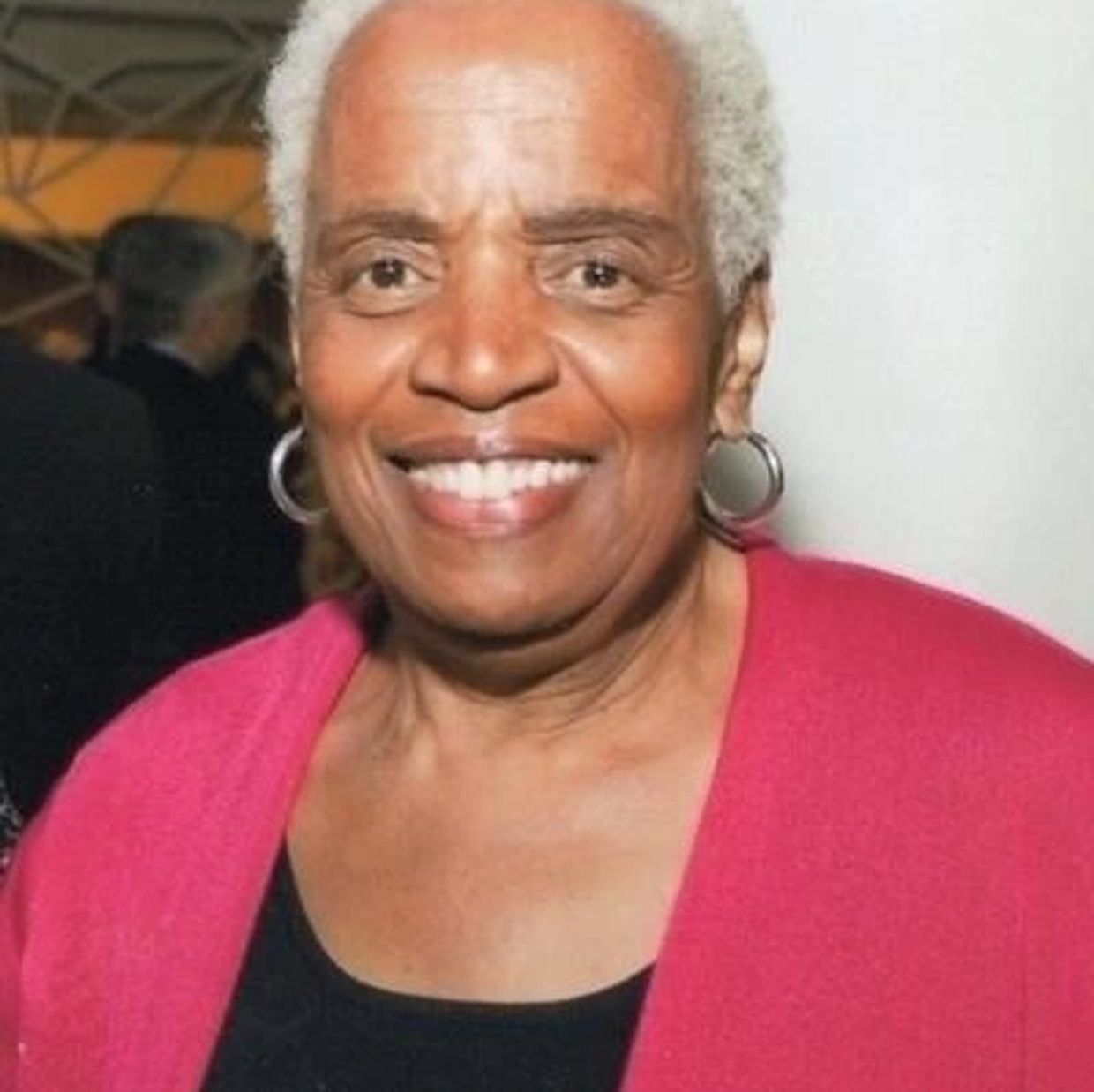 Smiling elderly woman with gray hair wearing hoop earrings and a red cardigan.