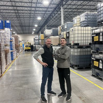 Two men standing and smiling inside a large warehouse filled with stacked containers.