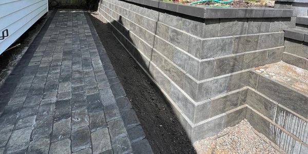 Narrow walkway with stone pavers and a retaining wall beside a house.