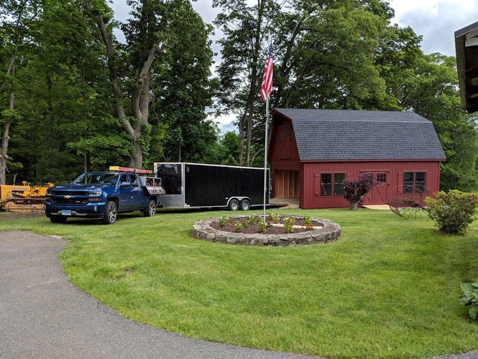 A blue truck with a trailer parked near a red barn and flagpole.
