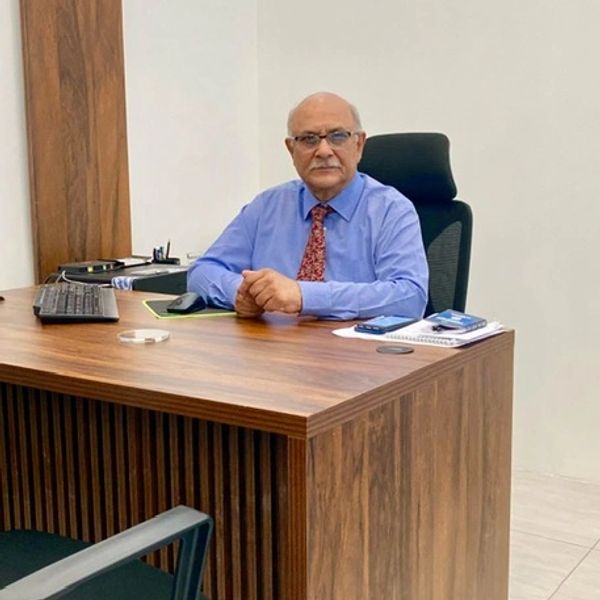 An elderly man in a blue shirt and red tie sitting at a wooden office desk.
