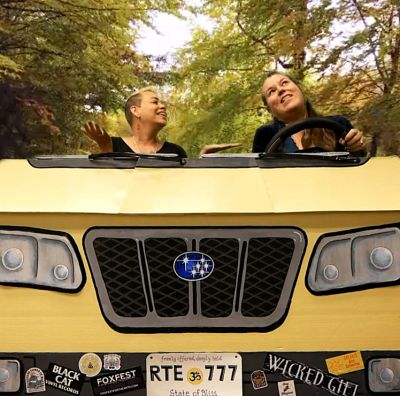 Two women happily driving a Subaru on a tree-lined road.