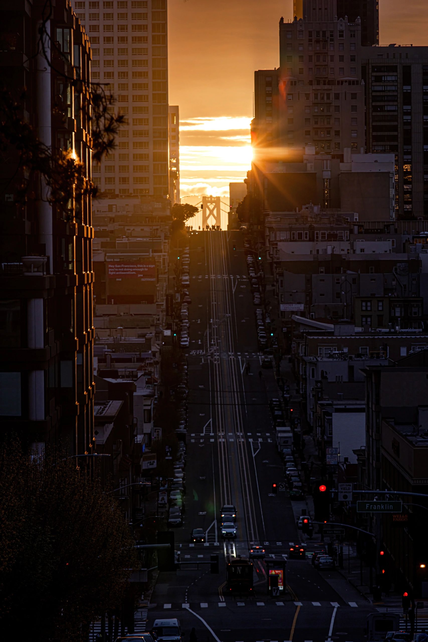 Sunset illuminates a city street with the Bay Bridge in the distance.