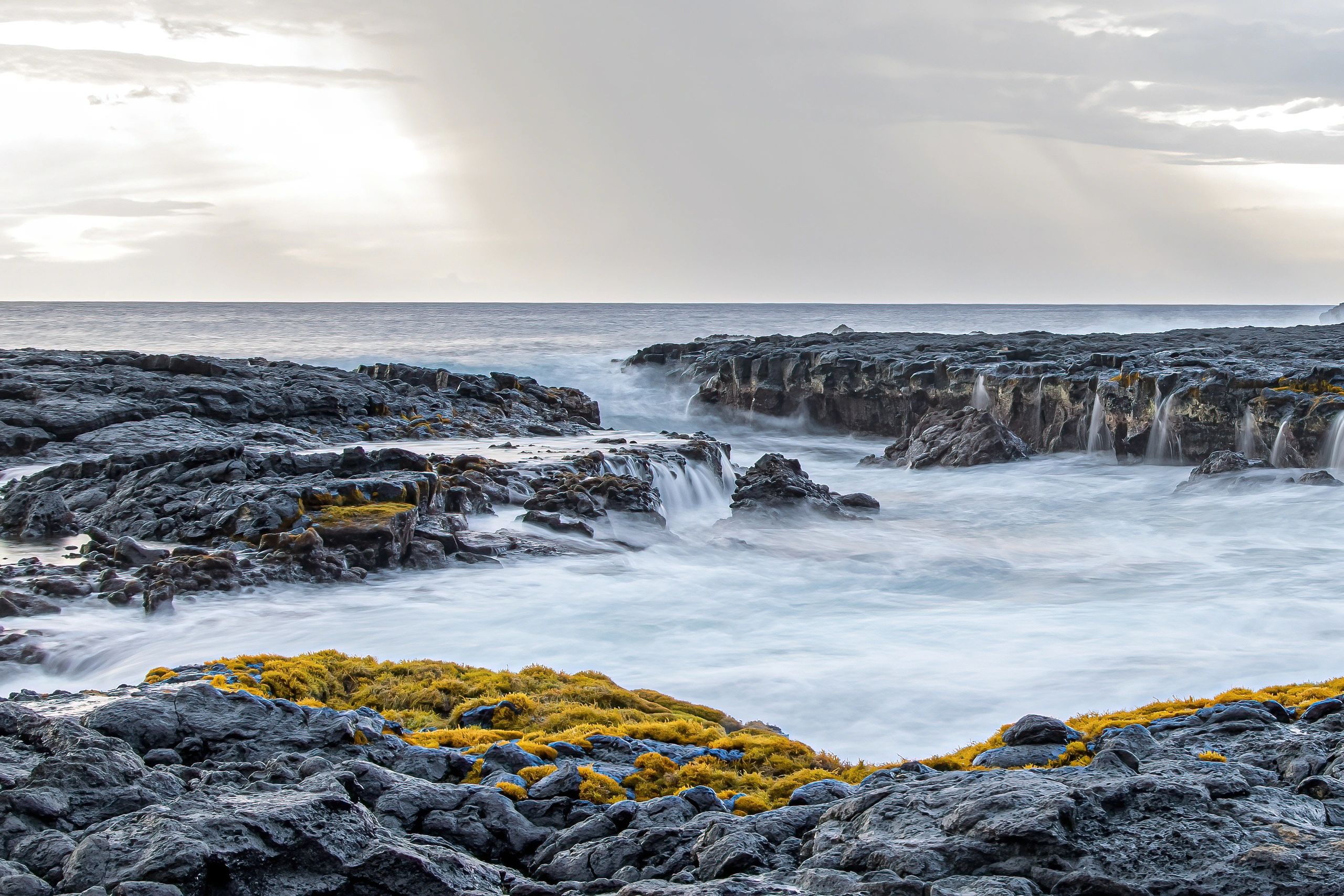 Rocky coastline with moss and gentle waves under a cloudy sky.
