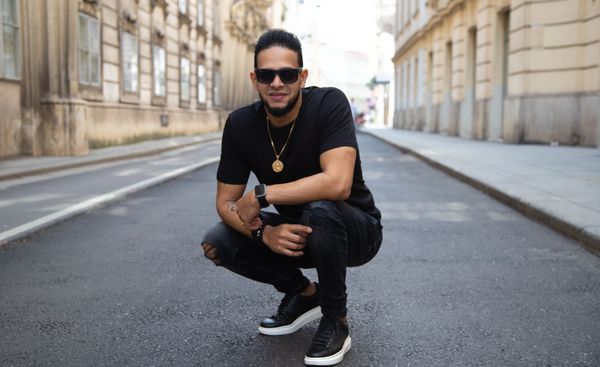 A man crouching on a city street wearing sunglasses and black clothes.