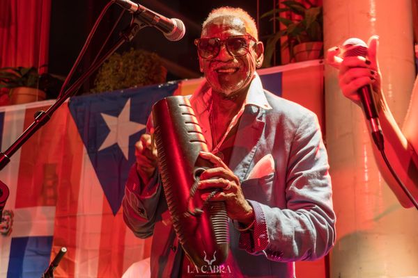 Musician playing a güiro on stage with a Puerto Rican flag background.