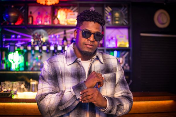 Man wearing sunglasses and a plaid jacket in a colorful bar setting.