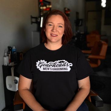 A woman in a black 'Gearheads Men's Grooming' t-shirt sitting in a barbershop chair.