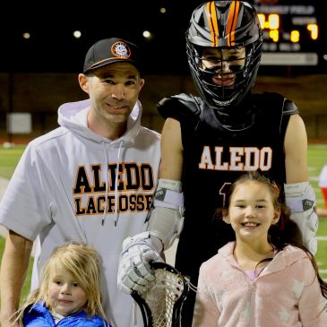 A lacrosse player and coach pose with two young girls on the field.