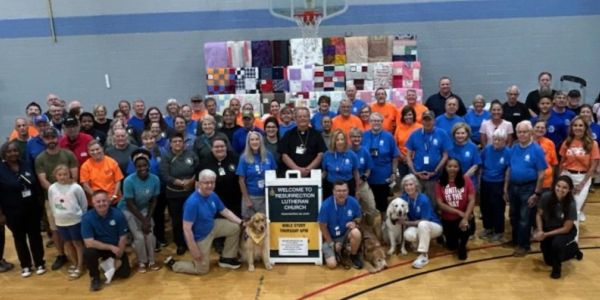 Large group of people with dogs posing in a gymnasium with colorful quilts in the background.