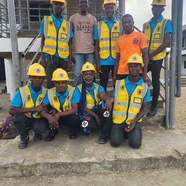 A group of young workers wearing yellow safety helmets and vests posing outdoors.
