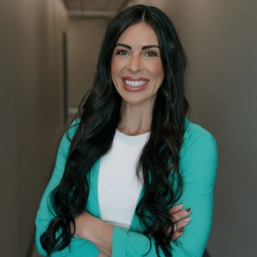 Younger dark haired woman posing and smiling for a photo. She is wearing a teal jacket with a white 