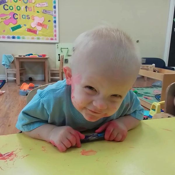 Toddler with paint on face and hands, smiling at a yellow table.
