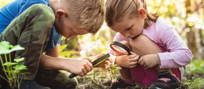 Two children examining a plant with a magnifying glass outdoors.