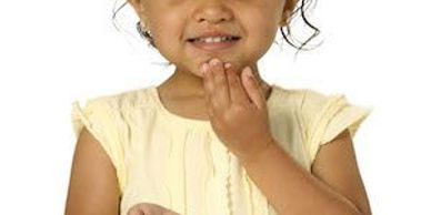 Smiling toddler with hand near mouth wearing a yellow dress.