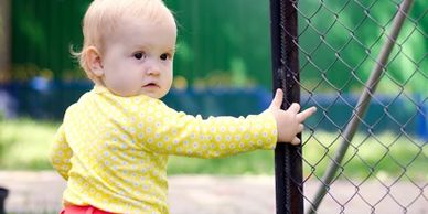 A toddler in a yellow shirt holding a metal fence outdoors.