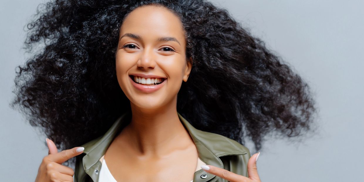 Happy woman with curly hair smiling and pointing at herself.