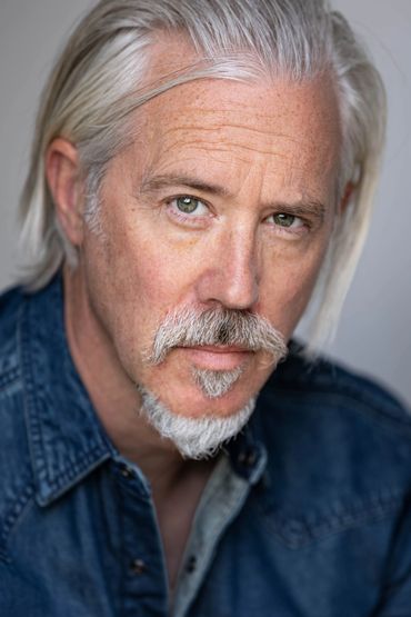 Close-up portrait of a mature man with gray hair and beard in a denim shirt.