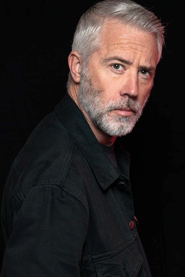Mature man with gray hair and beard in black jacket against black background.