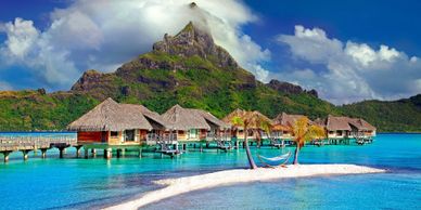 Overwater bungalows with a mountain backdrop and a hammock on a small sandy island.