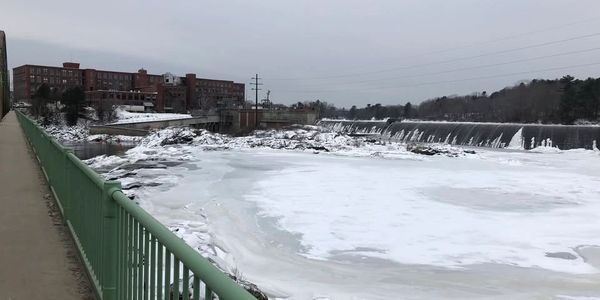 Frozen river with a dam and industrial buildings in the background.