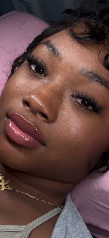 Close-up of a woman with glossy lips and long eyelashes lying on a pink pillow.