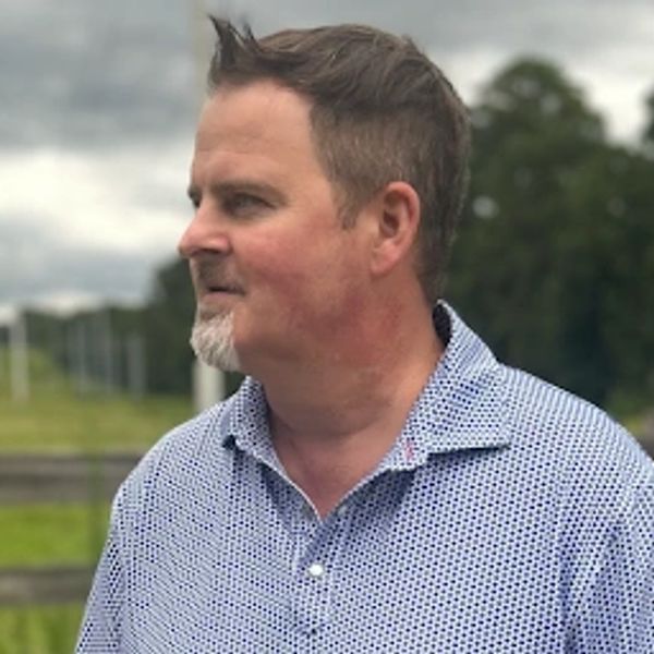 Man with short hair and a beard wearing a patterned shirt outdoors.