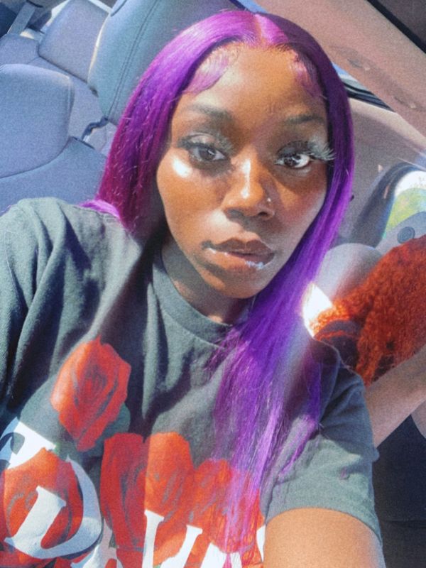 Young woman with vibrant purple hair sitting in a car, wearing a black shirt with red roses.