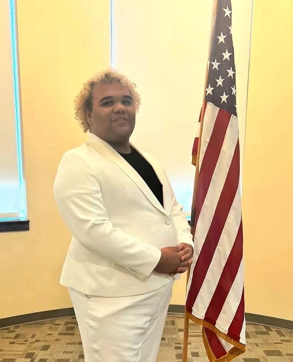 Man in white suit standing next to an American flag indoors.