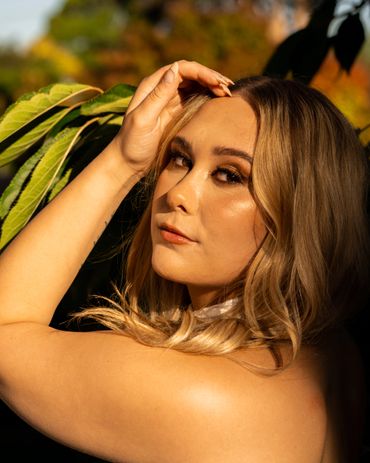 Sunlit portrait of a woman with blonde hair and natural makeup near green leaves.