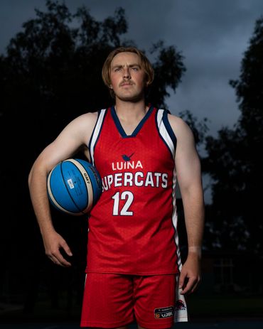 Basketball player in red Luina Supercats jersey holding a blue basketball outdoors.