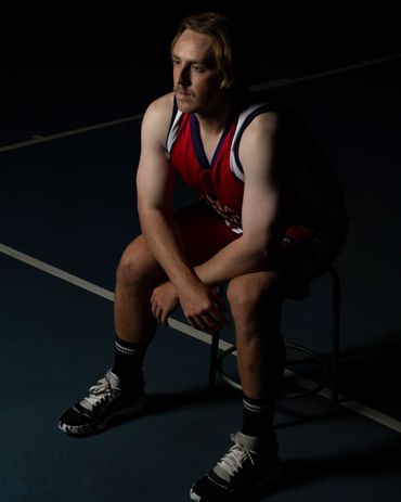 A basketball player in red jersey sits thoughtfully on a stool in a dark court.