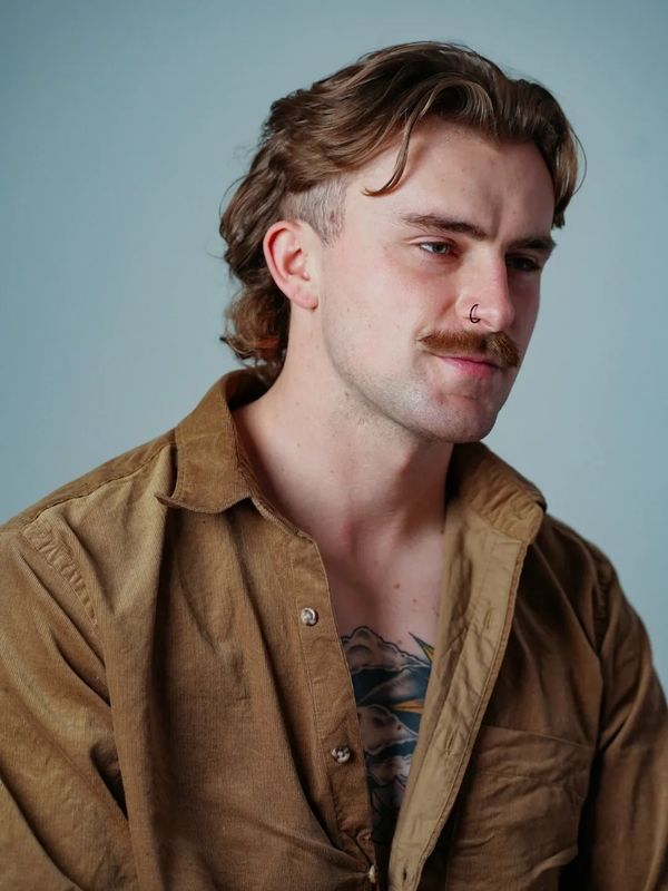 Man with a mustache, nose ring, and tattoo, wearing a brown shirt.