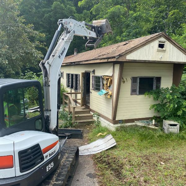 Bobcat excavator demolishing a small house in a green wooded area.